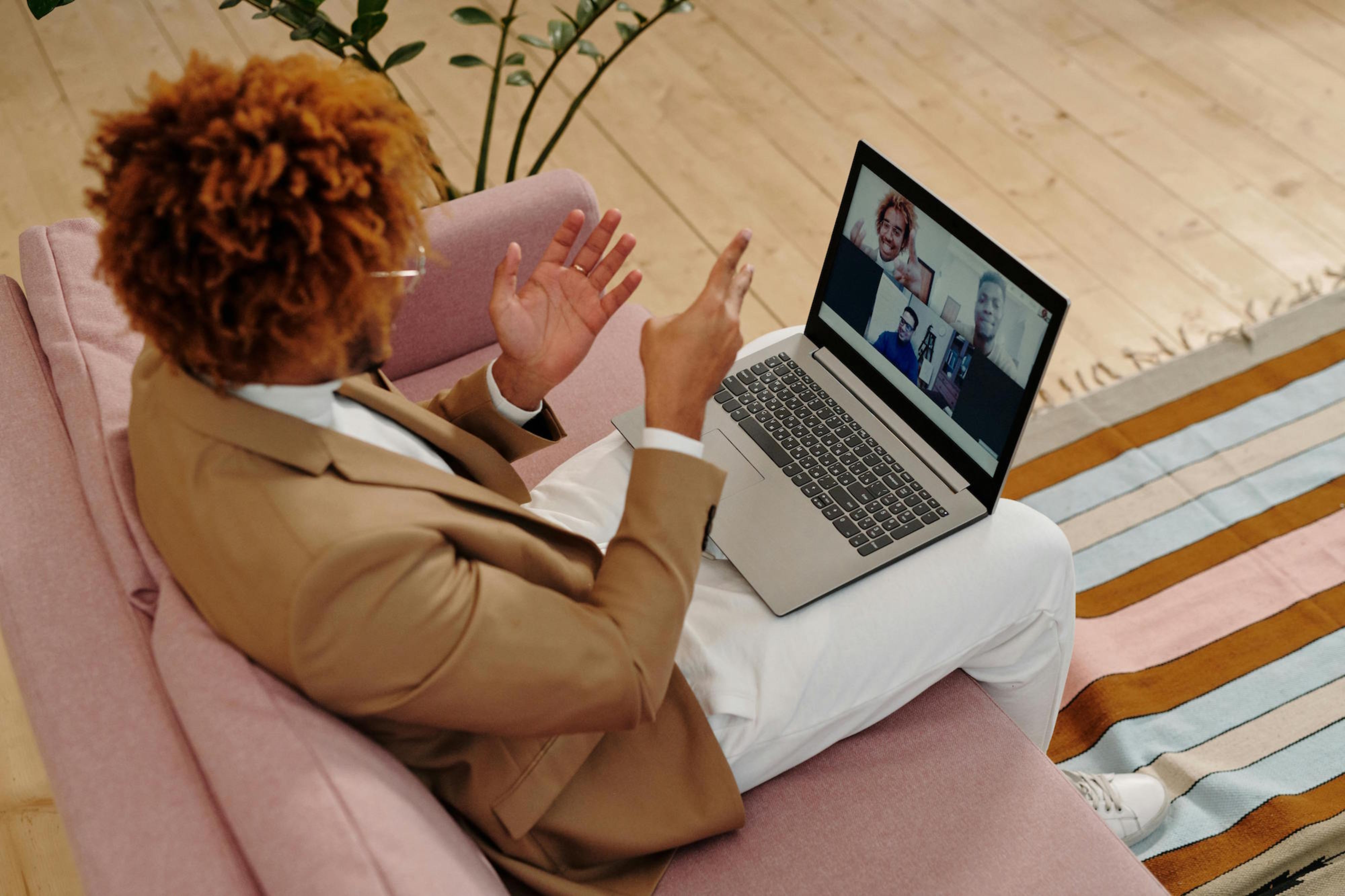 Person sitting on a pink chair using a laptop with a video call on the screen, in a room with wooden floor and striped rug.